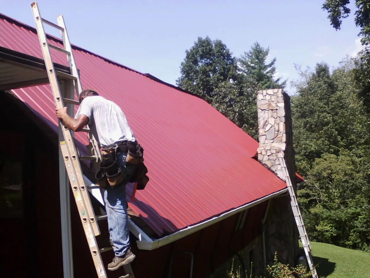 Roofer on a ladder beside a red metal roof and stone chimney — Soffit & Fascia Repair by Roofing Brothers in Wells